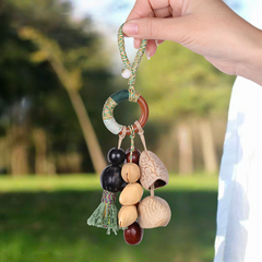 Handcrafted natural seed pod wind chime charm featuring a wooden ring wrapped in green thread, dangling various dark seeds, nuts, and a small green tassel, unique boho-style hanging accessory.