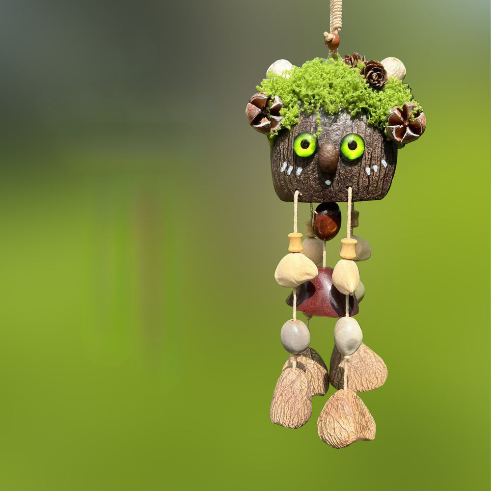 Handcrafted natural seed pod wind chime featuring a cute character shell with lush green mossy hair, large googly eyes, and a small red painted mouth, unique boho-style hanging home decor.