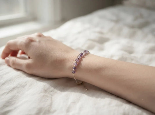 A close-up of a delicate amethyst crystal bracelet on a wrist, resting on soft, rumpled white linen bedsheets in gentle, natural morning light.