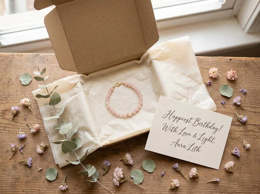Rose quartz crystal bracelet in an open gift box with dried flowers and a handwritten birthday card, warm natural light, feminine birthday gift scene.