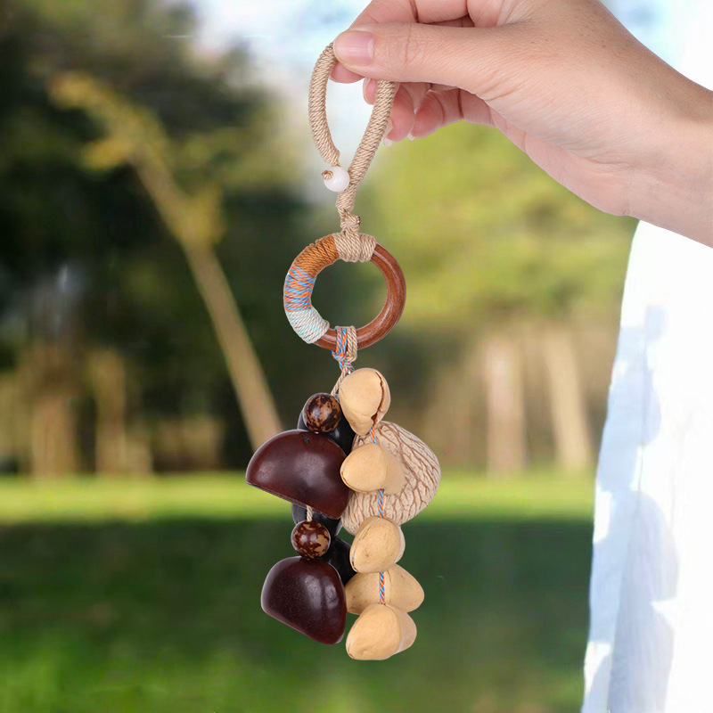 Handcrafted natural seed pod charm featuring a wooden ring wrapped in green thread, dangling various dark seeds, nuts, and a small green tassel, unique boho-style hanging accessory.