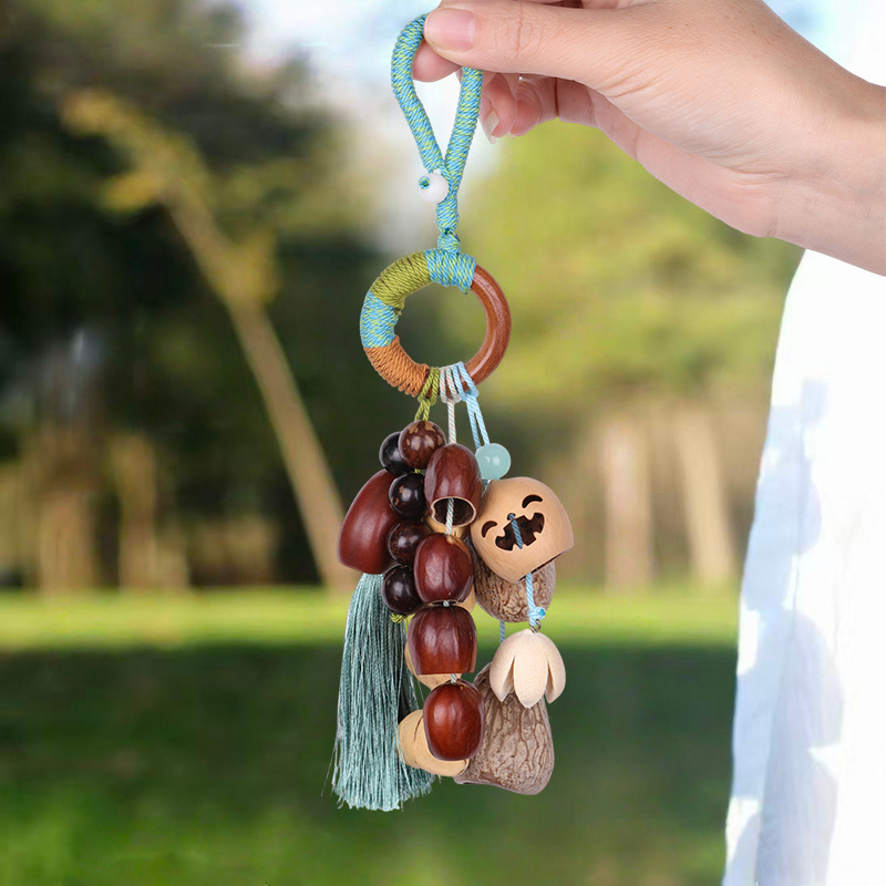 Handcrafted natural seed pod wind chime charm featuring a wooden ring wrapped in green thread, dangling various dark seeds, nuts, and a small green tassel, unique boho-style hanging accessory.