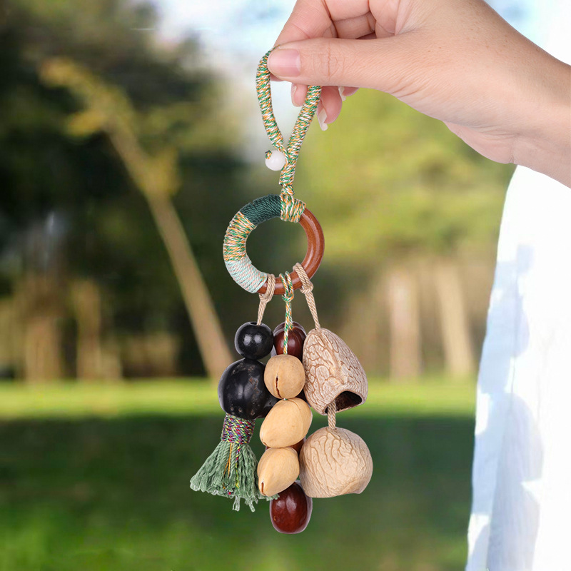 Handcrafted natural seed pod wind chime charm featuring a wooden ring wrapped in green thread, dangling various dark seeds, nuts, and a small green tassel, unique boho-style hanging accessory.